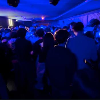 DJ in red shirt performing on stage with a lively crowd dancing under blue and white lights in a club setting.