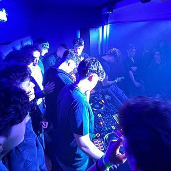 DJ in red shirt performing on stage with a lively crowd dancing under blue and white lights in a club setting.